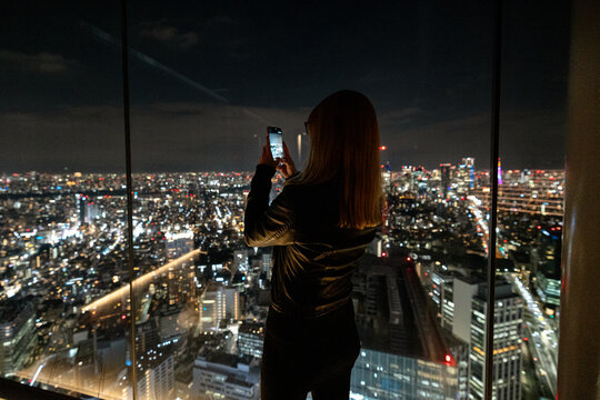 Tokyo, Japan - March 9, 2026: A woman photographing the nighttime views of traffic and city lights viewed from above Shibuya Scramble, Tokyo, Japan.

