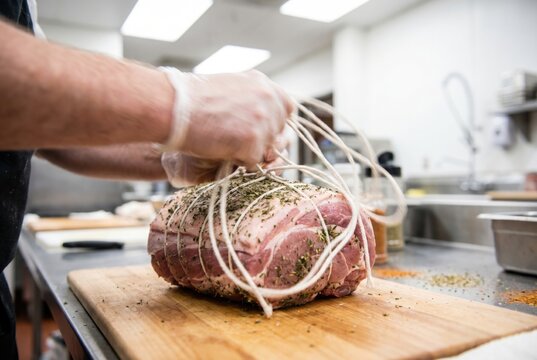 Butcher wearing clear gloves tightly pulling cotton twine to tie up a large seasoned raw pork roast on a wooden cutting board