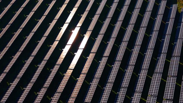 Geometric pattern of solar panels with intense sun glare. High angle shot of diagonal photovoltaic rows featuring strong light reflection on several modules.