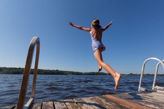 A thrilled child wearing goggles leaps from a wooden pier into sparkling blue lake water with outstretched arms on a sunny summer vacation day bursting with pure happiness and carefree holiday joy
