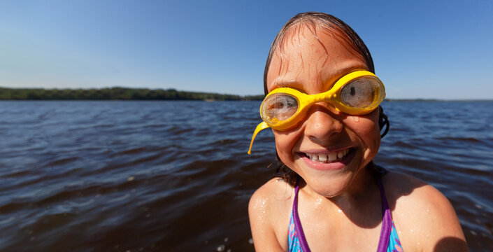Smiling child wearing yellow goggles enjoys swimming in lake with proper gear for young swimmers. Little girl plays cheerfully in water on summer day building skills and confidence. Kid learns proper.