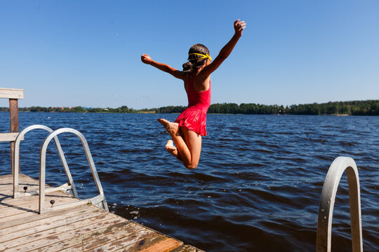 Daring child leaps off wooden dock into lake building confidence through exciting water sport. Brave little girl pushes her limits on day above water. Kid develops courage and athletic skill through.