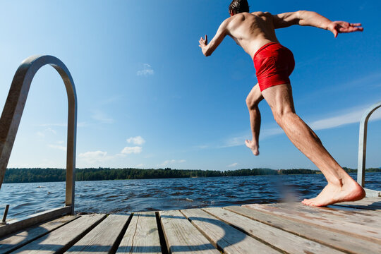 Man leaps off a dock into a lake on a sunny summer day. Guy in red swim trunks mid-air with outstretched arms in an athletic jump. Adult experiences thrilling outdoor recreation and refreshing swim