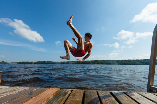 Man does backflip off wooden dock into lake water on sunny summer day. Guy in red trunks performs acrobatic flip midair with daring stunt outdoors. Spectacular dive carries risk of injury from pier