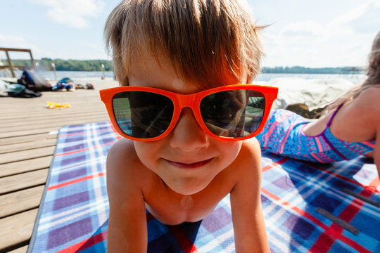 Happy boy relaxes on plaid blanket on wooden dock wearing red sunglasses and swimsuit beside calm lake. Child in warm summer day with carefree laid back mood. Kid enjoys leisure and outdoor peace