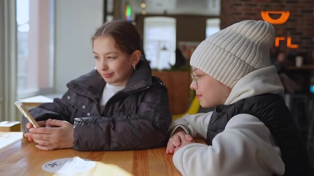Two girls in winter hats looking at phone and talking at cafe table in warm light. Phone, winter hats, cafe, talking, girls.