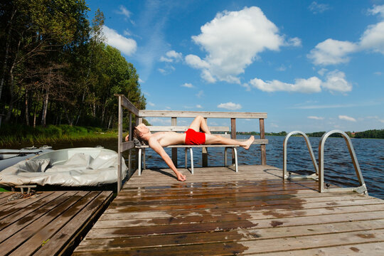 Man relaxes on a lounge chair at a wooden dock by a lake on a warm summer day. Guy in red tank top resting near calm water. Adult enjoys peaceful outdoor leisure and serene vacation