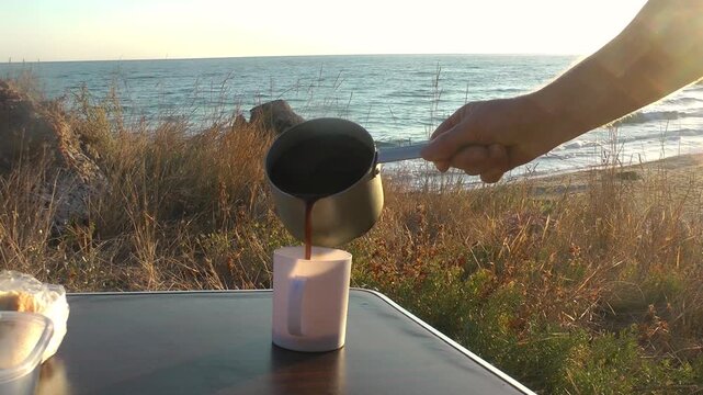 A hand pours hot coffee from a cezve into a cup on a seaside table. Morning coffee, coastal travel, beverage relaxation.