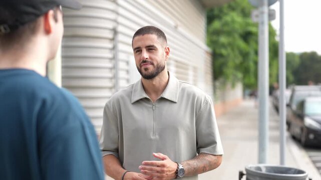 Man stands outside, talking to another person. He wears a light polo shirt and shows calm focus. The scene happens on a quiet sidewalk near buildings. One man faces the camera, appearing engaged