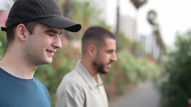 Two men walk side by side on a city sidewalk. One man wears a cap, smiling gently. The other walks beside him, focused ahead. Soft greenery lines their path behind them. Urban scene captures casual