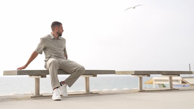 Man sits calmly on bench by ocean. He looks thoughtful, relaxed, slightly tired. Bird glides above in distant sky. Peaceful beachside scene, quiet, contemplative. Ideal for wellness, travel