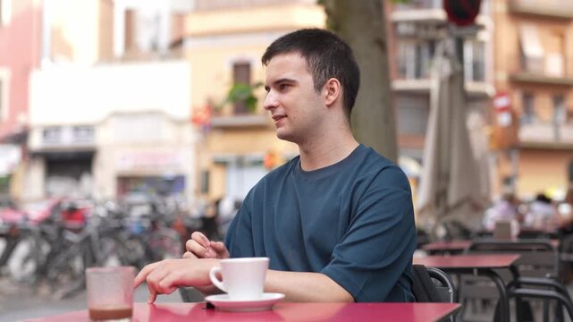 Young man ponders at outdoor cafe table. He rests chin on hand, gazing thoughtfully. Soft-focus background reveals city streets and parked bikes. Cafe scene evokes calm, quiet moments of reflection