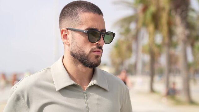 Man with short hair and sunglasses walks along a beach. He wears a light-colored shirt, looks thoughtful, and gazes off-camera. Palm trees and distant figures add beach ambiance
