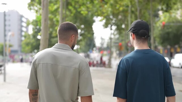Two men walk side by side down a tree-lined street. One wears a beige shirt, the other a dark t-shirt and cap. They move casually, heads slightly lowered. The scene feels relaxed, unhurried