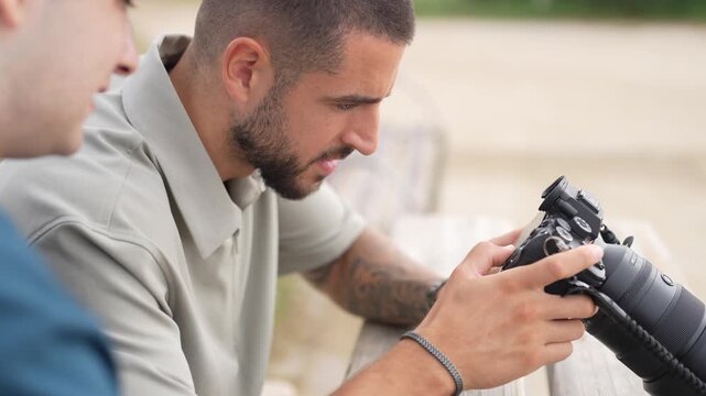 Two men examine a camera together. One man focuses intently on the lens. The other watches closely, curious. They sit near a wooden bench outdoors. Perfect for photography or travel storytelling