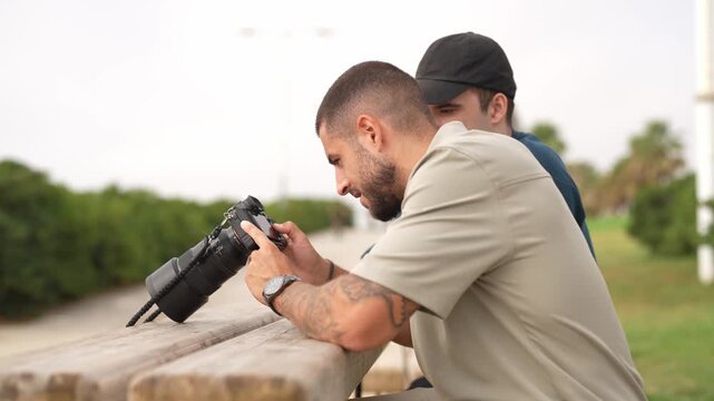 Man focuses on camera lens, capturing moment. Second man watches closely, ready to assist. Outdoor setting, natural light, calm atmosphere. Ideal for travel, lifestyle, or documentary projects