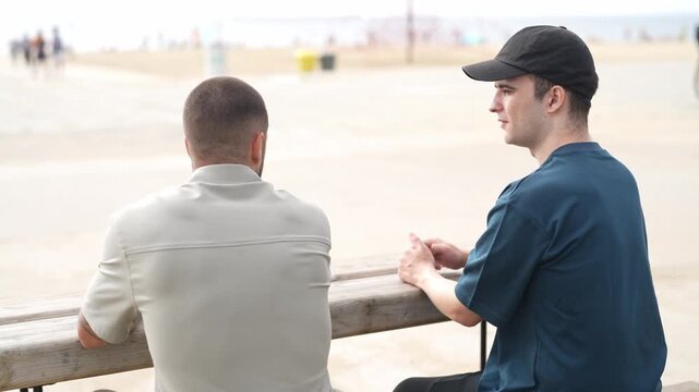 Two men sit on a beach bench, watching the ocean. One wears a cap, the other a light shirt. They appear calm, focused on the horizon. The beach is quiet, with distant figures in the background