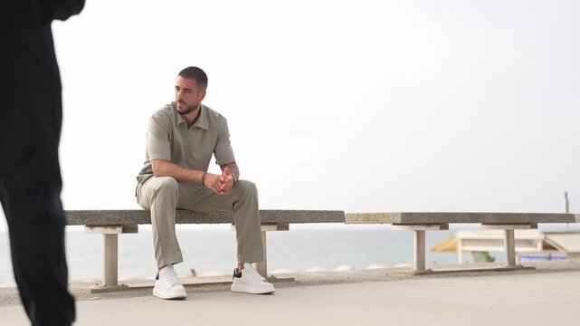 Man sits calmly on a bench near the sea. Two people walk past him, unaware of his presence. Bright daylight highlights the scene. The man appears relaxed and thoughtful