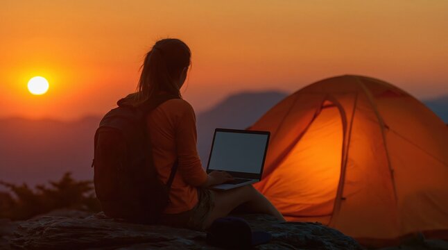 Person sits with laptop next to glowing tent, enjoying beautiful sunset in remote outdoor location. High quality photo