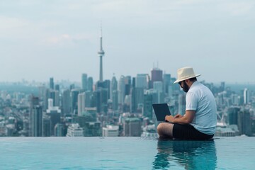 Naklejka premium Man using laptop by infinity pool overlooking Toronto skyline and CN Tower. Digital nomad working remotely with freedom to travel