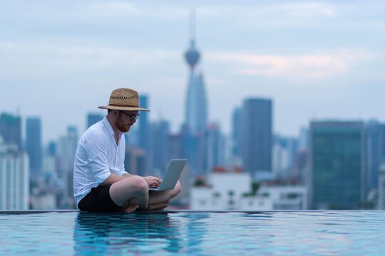 Young man using laptop while sitting on the edge of a rooftop infinity pool, enjoying remote work with city skyline view