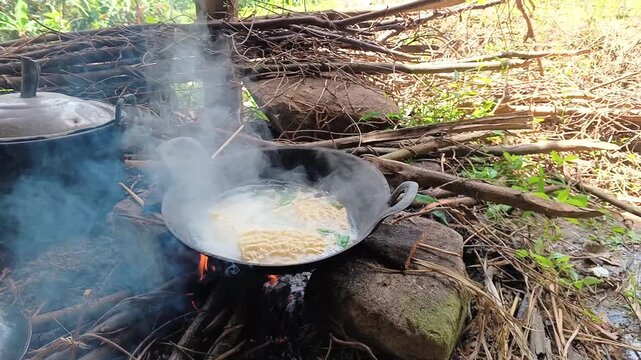 Traditional outdoor cooking of noodles on a wood fire, person stirring mie in a metal wok over an open flame.