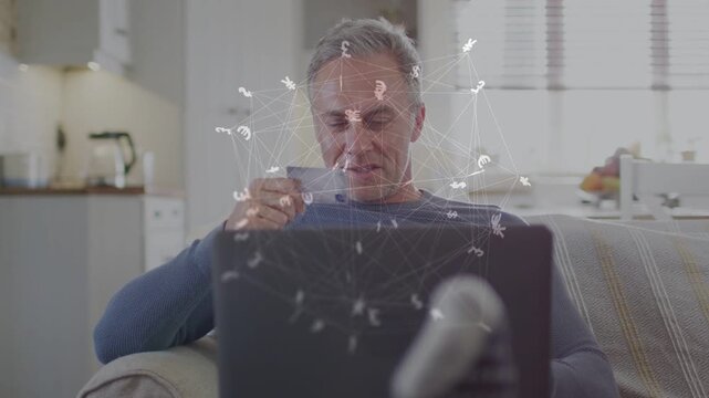 Mature man sipping cup, browsing laptop from sofa causing money icons orbiting head, checking funds