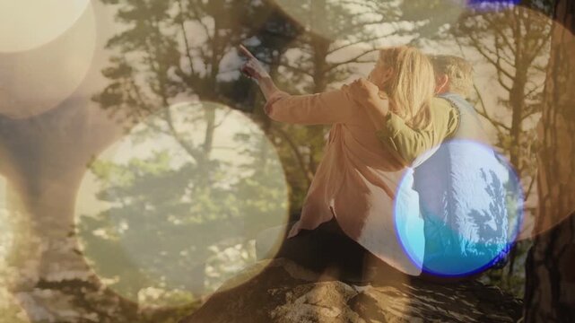 Couple sitting rocky outcrop, woman pointing, bokeh drifting over them, showing valley for travel