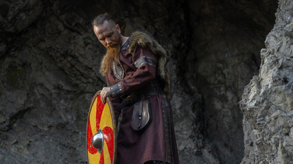 Viking warrior stands confidently against a rocky backdrop, clutching his shield and adorned with fur and leather. © horimono