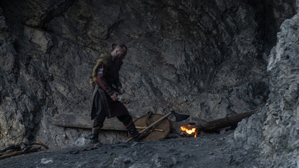 A rugged Viking stands near a campfire within a rocky cave, sharpening his axe. © horimono