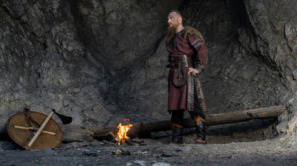 A rugged Viking warrior stands confidently amidst a rocky, dimly lit cave with his shield and axe nearby. © horimono