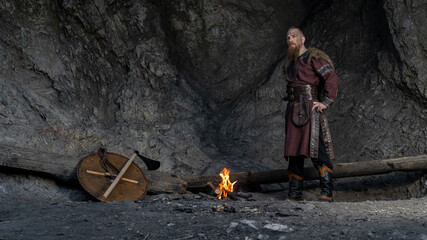 A rugged Viking warrior stands near a campfire, axe and shield resting nearby within a rocky cave. © horimono