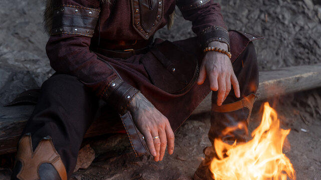 A man dressed as a Viking sits near a campfire, his hands resting on his tunic and boots.