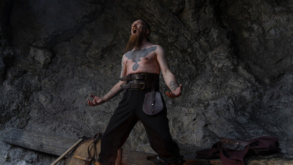 A muscular, bearded man with tattoos stands dramatically amidst a rocky cavern, arms outstretched and mouth open. © horimono