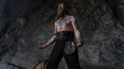 A muscular, bearded man with tattoos stands dramatically against a rocky backdrop, arms raised and mouth open in a shout. © horimono