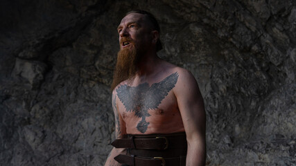 Tattooed man with long beard stands shirtless against rocky backdrop, looking up with an expression of awe. © horimono