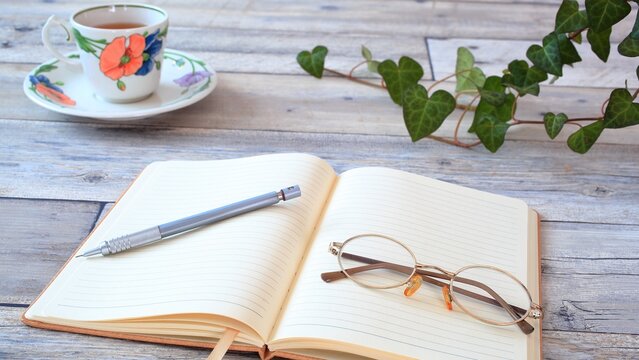 Open notebook with pen and eyeglasses on wooden table, tea cup and green plant, cozy lifestyle workspace