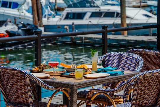 Outdoor dining on a marina with food and drinks near boats. People enjoy food and drinks at a table by water with boats in the background.