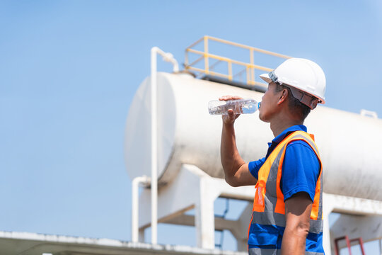 Thirsty engineer taking a break and drinking from water bottle at oil refinery industrial site, Asian technician refreshing with bottled water during outdoor work at fuel storage tank