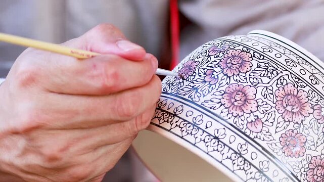 Close up of artisan hand painting intricate floral patterns on ceramic bowl with fine brush.