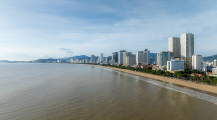 Obraz premium A panoramic view of the Nha Trang beach in Vietnam, with its sandy shore, palm trees, and modern high-rise buildings lining the coast. The sea is calm under a partly cloudy sky.