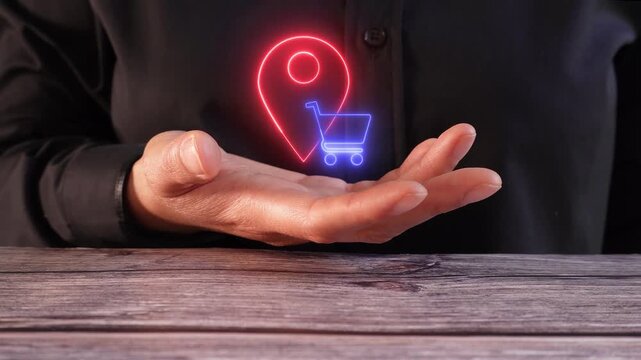 Location-based shopping. Close-up of a woman's hand at a table holding a virtual location pin icon and a shopping cart.