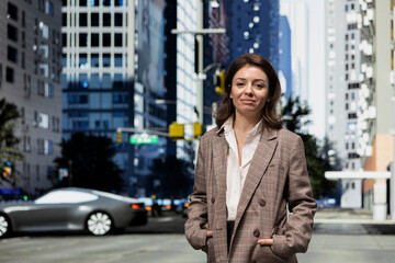 Fototapeta premium Portrait of smiling woman walking past modern office buildings in city financial hub. Happy citizen with hands in pockets commuting on foot through developed metropolitan infrastructure