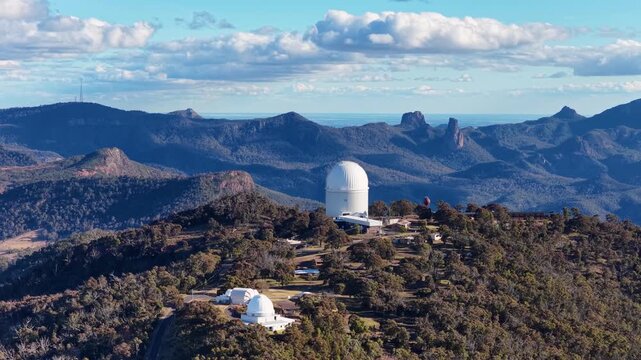 Siding Spring Observatory drone footage showing white telescope domes on mountain ridge against blue sky and clouds in New South Wales.