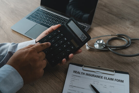 Hand using calculator on medical insurance claim form beside laptop and stethoscope performing financial calculation for healthcare expense budgeting, accounting report analysis and cost management pr