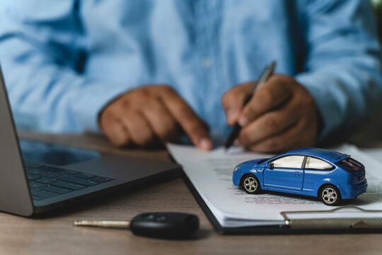 Hand signing car finance loan document form with laptop and blue toy car model on desk with key, representing online auto financing application, digital car loan approval process, remote vehicle finan