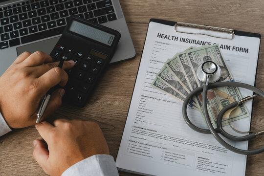 A healthcare professional uses a calculator to carefully process health insurance claim reimbursement costs, with a stethoscope and US dollar bills placed on a medical claim form clipboard.