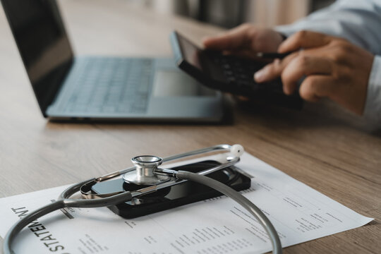 A doctor carefully uses a calculator and laptop to review a medical billing statement for insurance cost analysis, with a stethoscope and smartphone resting on the document on a wooden office desk.
