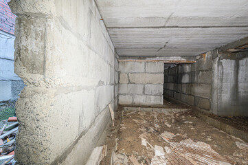 An unfinished concrete room with rough block walls, uneven dirt floor, scattered construction debris, exposed rebar, dim lighting, unsealed ceiling, signs of moisture © evgeniykleymenov