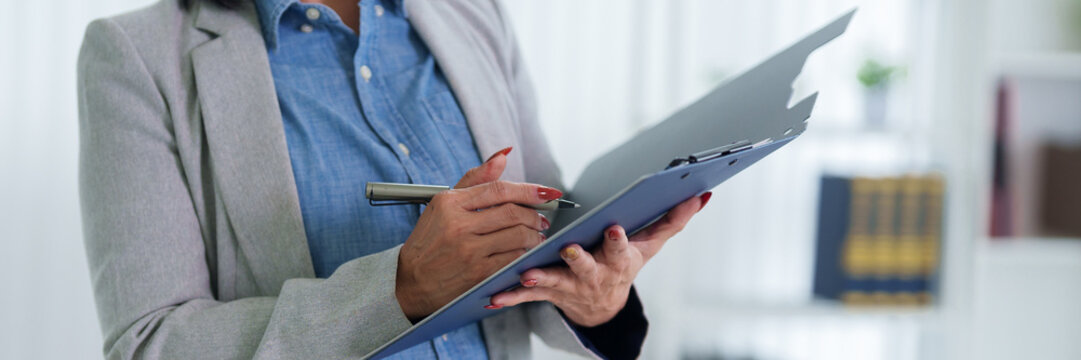 businesswoman professional writing notes document clipboard paperwork reviewing report office hands formal business suit gray blazer blue shirt corporate